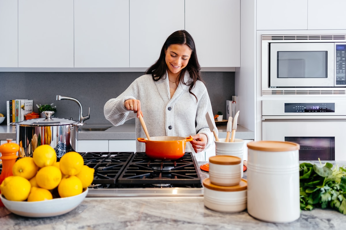 Woman in a grey cardigan stirring an orange pot with a wooden spoon on a gas stove, with lemons and kale on a marble kitchen island