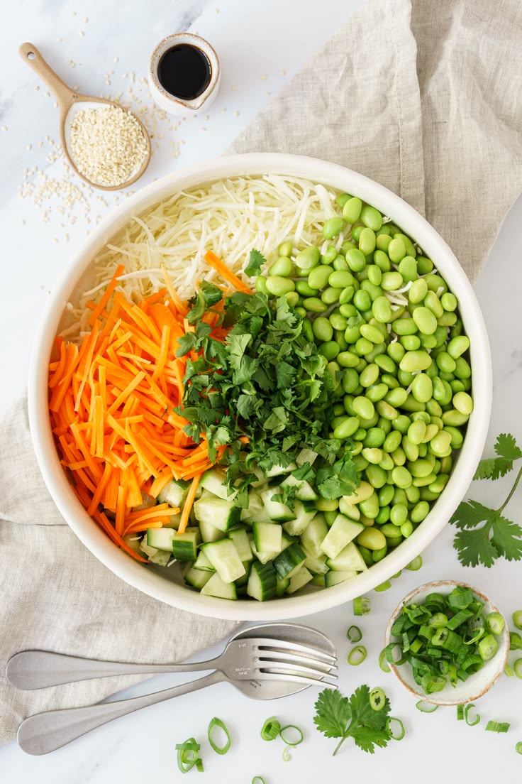 Overhead photo of a white bowl with shredded cabbage, julienned carrots, edamame, diced cucumber, and cilantro on a marble counter